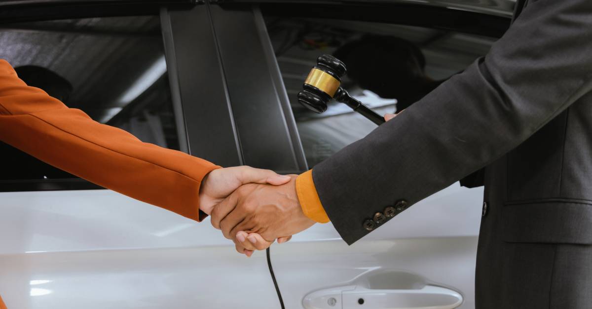 A close-up of two people wearing suits shaking hands in front of a vehicle door. The right person holds a gavel.