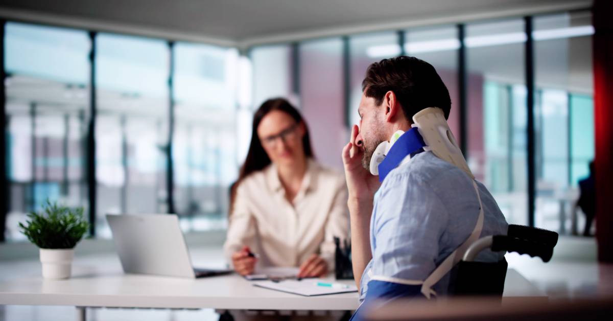 A man in a neck brace sits across the table from a woman in a white shirt and glasses. She has a clipboard for him to fill out.