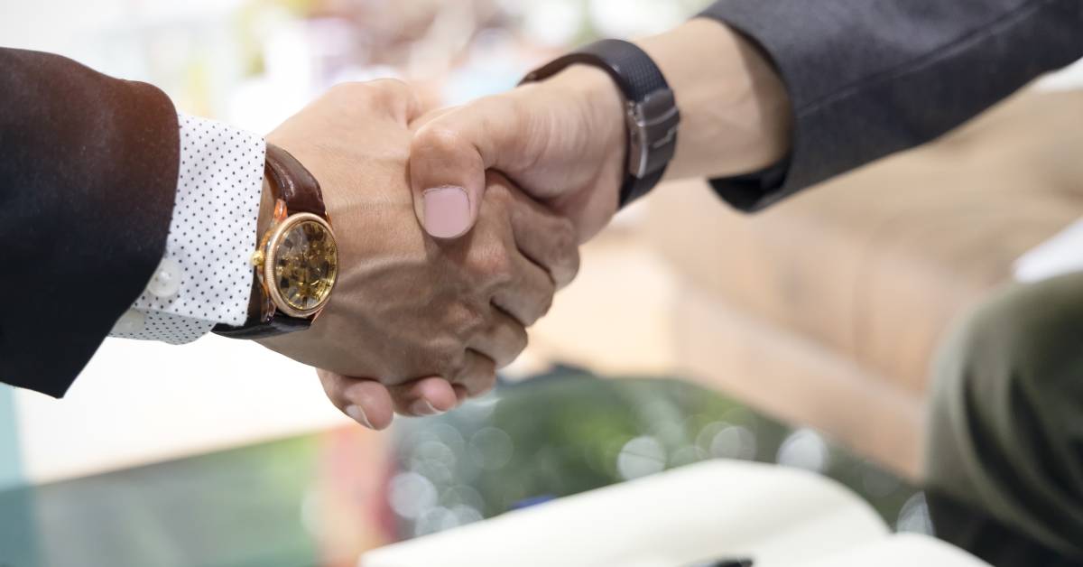 A close-up of two men shaking hands. One man wears a gray suit and the other wears a black suit, while both of them wear watches.