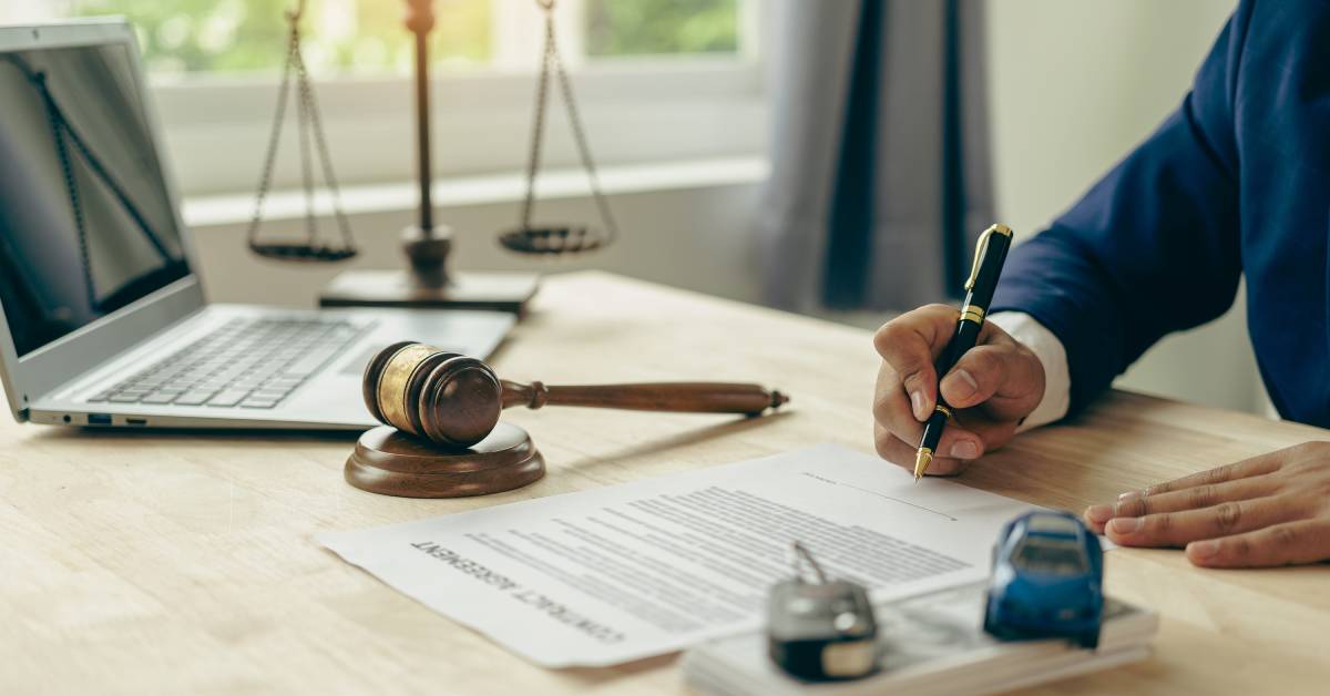 A man in a blue suit sitting at a desk, signing a form. A toy car, car key, laptop, scales, and gavel also rest on the desk.