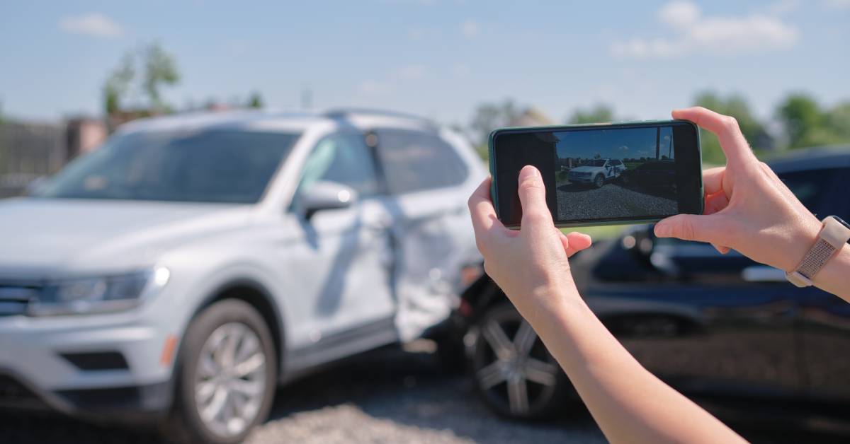 A person's hands holding a phone from off-screen, taking pictures of two cars after an accident in the background.