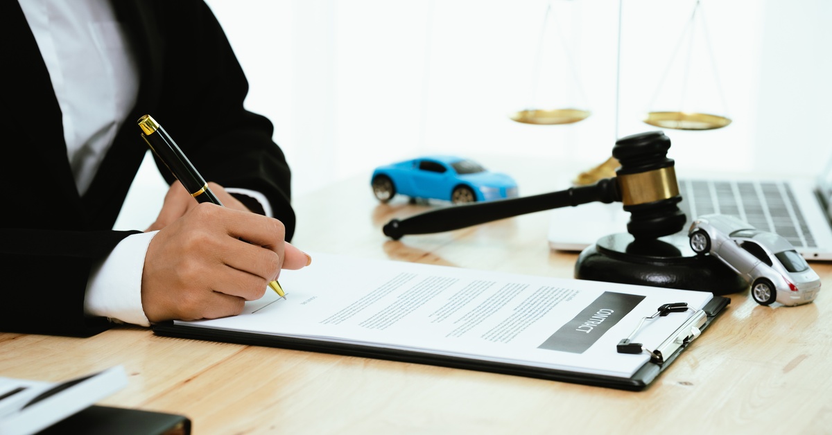 A close-up of a man wearing a black suit sitting at a desk with a pen and writing on a clipboard. A gavel sits on the desk.