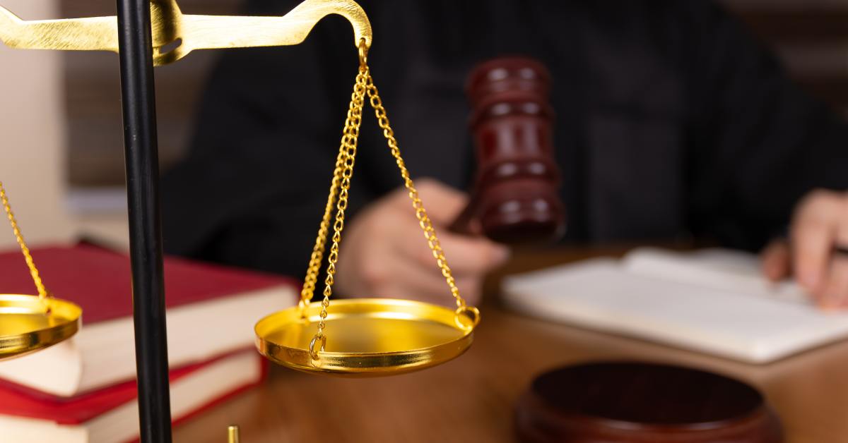 A close-up of a set of golden scales sitting on a judge's desk with a judge holding a gavel in the background.
