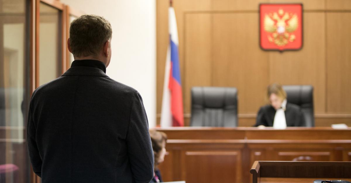 A man standing in a courtroom with his back to the camera, looking out at a judge's podium, a flag, and a symbol on the wall.